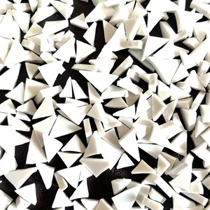 May include: A collection of small, irregularly shaped white glass mosaic tiles scattered across a dark surface. Each piece features sharp, angular edges and a smooth, opaque finish, creating a high-contrast pattern of white shards against the black.