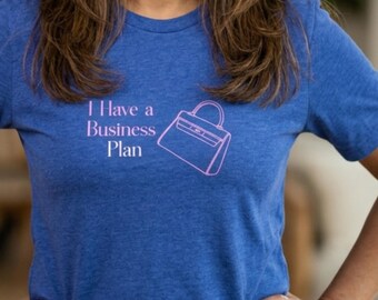 May include: A heathered royal blue short-sleeve t-shirt featuring the light pink text "I Have a Business Plan" printed on the chest, accompanied by a simple light pink line-art graphic of a structured handbag positioned to the right of the text.