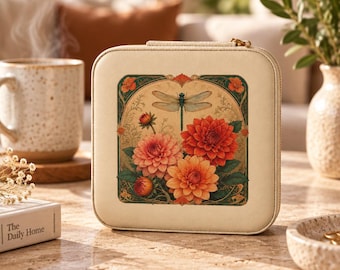 May include: A square, cream-colored jewelry case featuring a central graphic print of a dragonfly hovering above a cluster of orange and red dahlia flowers, framed by a decorative border with green and gold accents and a gold zipper closure.