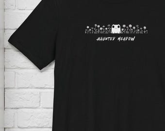 May include: A black short-sleeved t-shirt featuring a white graphic print of a small creature surrounded by flowers and the text "HAUNTED MEADOW" centered on the chest. The design is rendered in a simple line art style on the front of the garment.