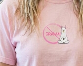 May include: A light pink t-shirt featuring a graphic of a white llama sitting in a lotus position. A pink circle with a diagonal line crosses out the word DRAMA, which is printed in pink capital letters next to the llama illustration.
