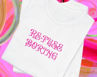 May include: A white short-sleeved t-shirt featuring the text REFUSE BORING printed in a stylized, decorative pink font across the chest. The garment is folded and presented against a vibrant, abstract background of swirling pink and orange hues.