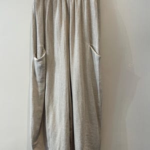 May include: Light beige linen trousers with an elasticated waistband and two large front pockets. The trousers have a relaxed fit and a slightly gathered, rounded hem. The fabric has a subtle textured appearance, hanging from a metal hanger.