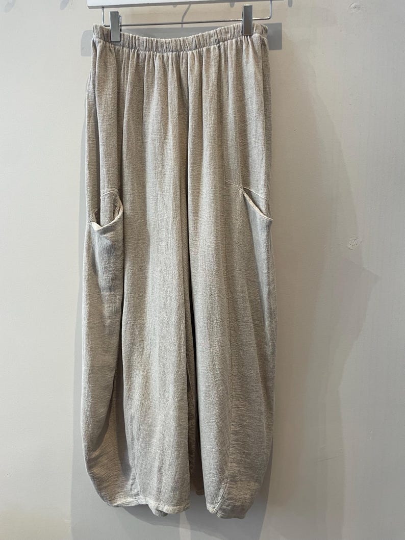 May include: Light beige linen trousers with an elasticated waistband and two large front pockets. The trousers have a relaxed fit and a slightly gathered, rounded hem. The fabric has a subtle textured appearance, hanging from a metal hanger.