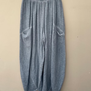 May include: Pale blue linen trousers with two large front pockets. The trousers have a relaxed fit and an elasticated waistband. The fabric has a textured appearance. The trousers are hanging on a white hanger.