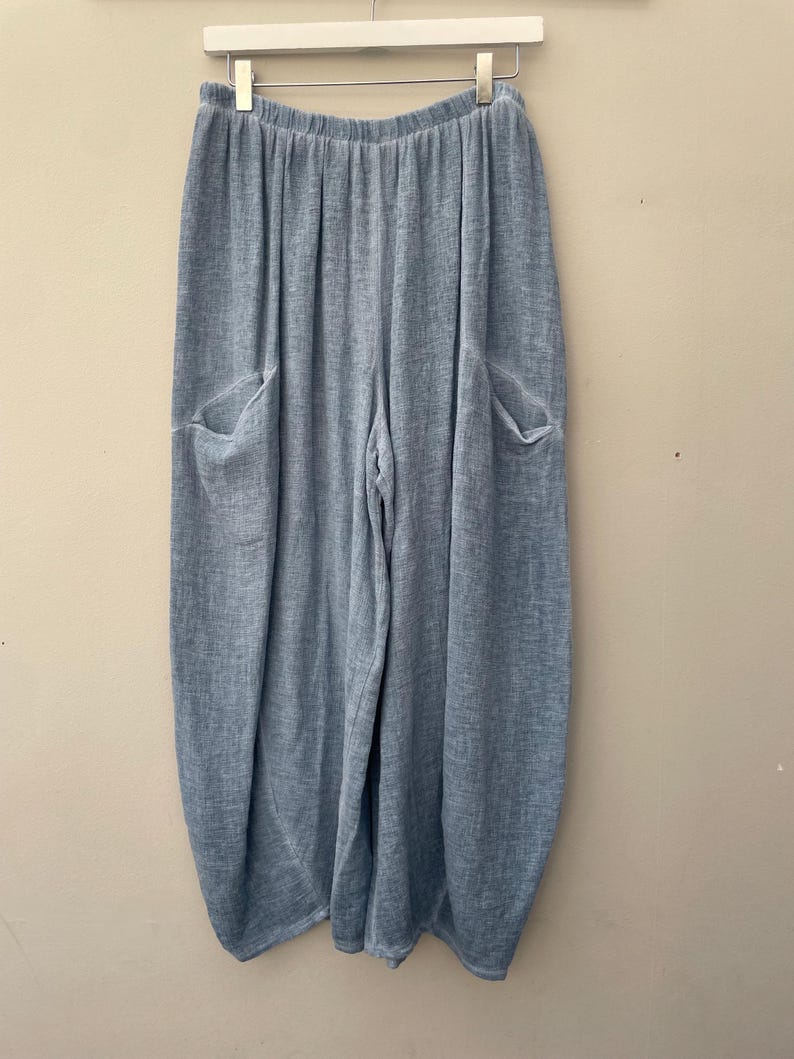 May include: Pale blue linen trousers with two large front pockets. The trousers have a relaxed fit and an elasticated waistband. The fabric has a textured appearance. The trousers are hanging on a white hanger.