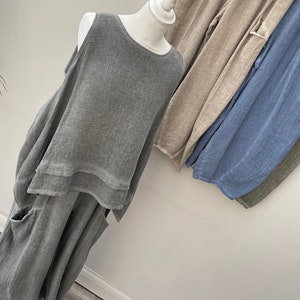 May include: A grey linen outfit on a mannequin, comprising a sleeveless top and wide-leg trousers with pockets. Additional linen trousers in beige, blue, and olive green are visible. The outfit has a relaxed, flowing design, suitable for warm weather.