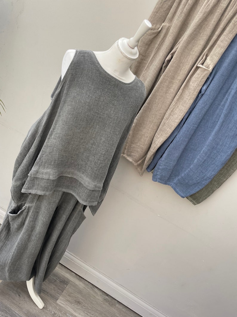 May include: A grey linen outfit on a mannequin, comprising a sleeveless top and wide-leg trousers with pockets. Additional linen trousers in beige, blue, and olive green are visible. The outfit has a relaxed, flowing design, suitable for warm weather.