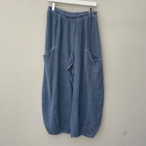 May include: Pale blue linen trousers with large side pockets, hanging from a white hanger. The trousers have a relaxed, wide-leg design and a gathered elastic waistband. The fabric has a textured appearance.