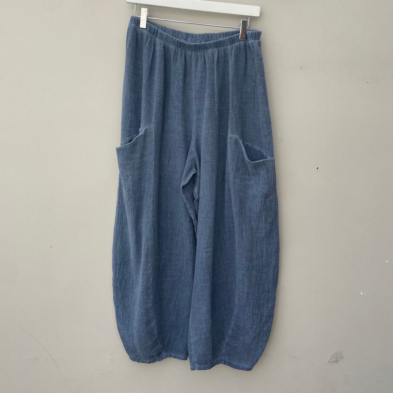 May include: Pale blue linen trousers with large side pockets, hanging from a white hanger. The trousers have a relaxed, wide-leg design and a gathered elastic waistband. The fabric has a textured appearance.