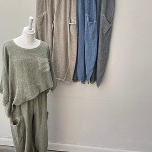 May include: A green linen outfit on a mannequin, with a matching top and trousers. Behind it, three pairs of linen trousers in beige, blue, and grey hang on a rail. Each pair has large pockets.
