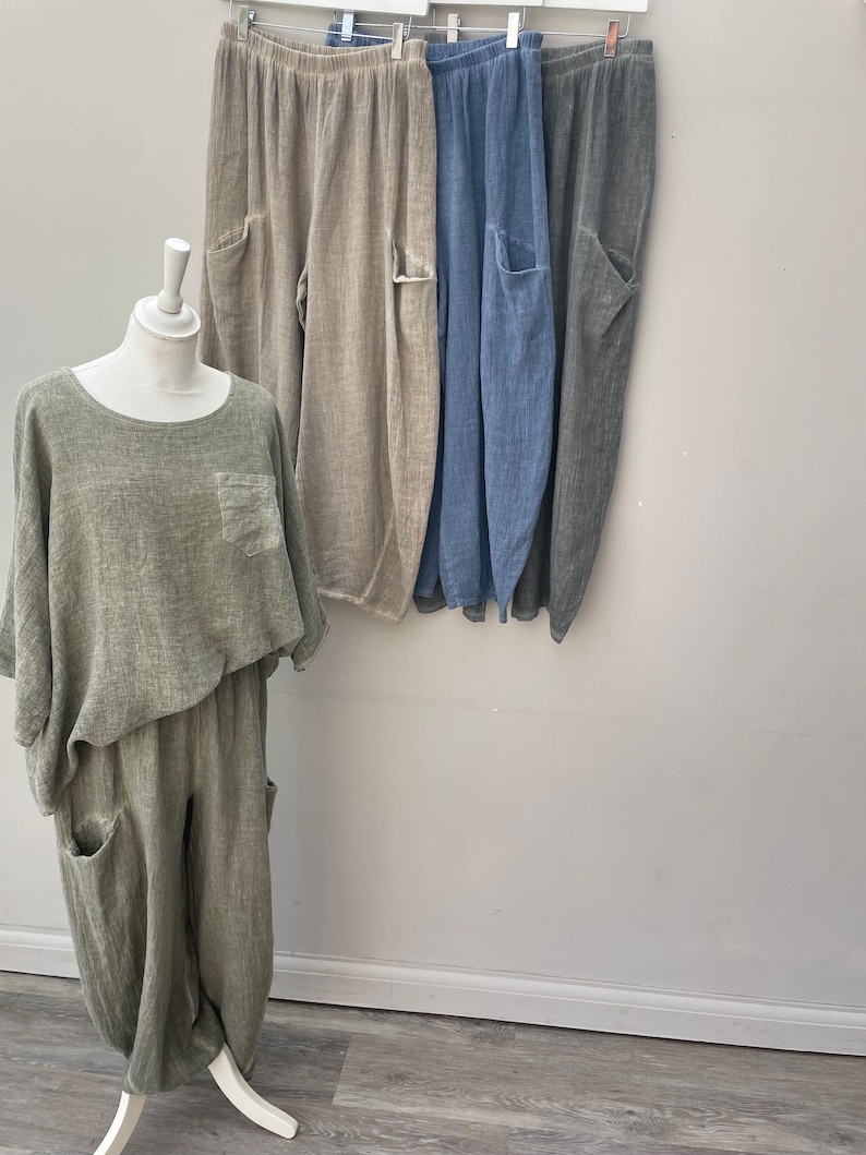 May include: A green linen outfit on a mannequin, with a matching top and trousers. Behind it, three pairs of linen trousers in beige, blue, and grey hang on a rail. Each pair has large pockets.