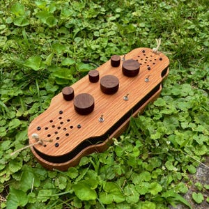 May include: A wooden electronic device with a light-colored top surface and dark-colored sides. The top features five circular dark wood knobs, several small metal toggle switches, and two perforated speaker-like areas with small black screws.