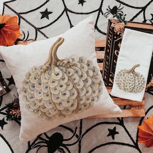 May include: A square off-white throw pillow and a rectangular white waffle-weave kitchen towel, both featuring a printed illustration of a pumpkin composed of layered oyster shells in shades of beige, cream, and soft blue-gray.