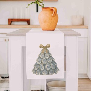 May include: A white table runner featuring a central graphic of a Christmas tree constructed from layered oyster shells. The tree is topped with a tan-colored bow. The design is rendered in shades of blue, gray, and tan, centered on the fabric.