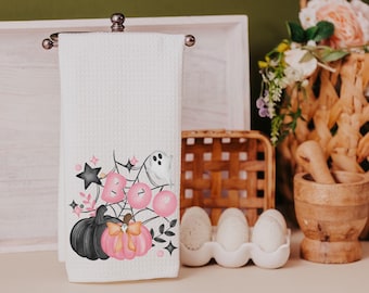 May include: A white kitchen towel with a pink and black Halloween design featuring pumpkins, a ghost, and the word "BOO" on a spiderweb.