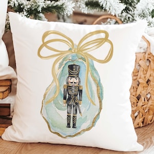 May include: A white square throw pillow featuring a centered watercolor illustration of a nutcracker standing inside a light blue and green oyster shell, topped with a large gold ribbon bow with gold-painted edges on the shell.