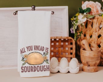 May include: White kitchen towel with a brown and white illustration of a loaf of sourdough bread and the text "All you knead is sourdough".