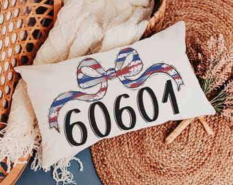 Op de afbeelding: Crèmekleurig rechthoekig kussen met een strikdesign in rood, wit en blauw gestreept, met het nummer "60601" in zwarte letters. Het kussen ligt op een geweven oppervlak. Het ontwerp doet denken aan een vlag.