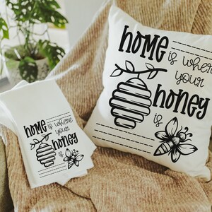 May include: A white throw pillow and a white kitchen towel with black text and illustrations. The pillow and towel both say "Home is where your honey is" with a bee and honeycomb design.