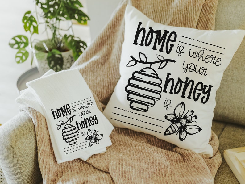 May include: A white throw pillow and a white kitchen towel with black text and illustrations. The pillow and towel both say "Home is where your honey is" with a bee and honeycomb design.
