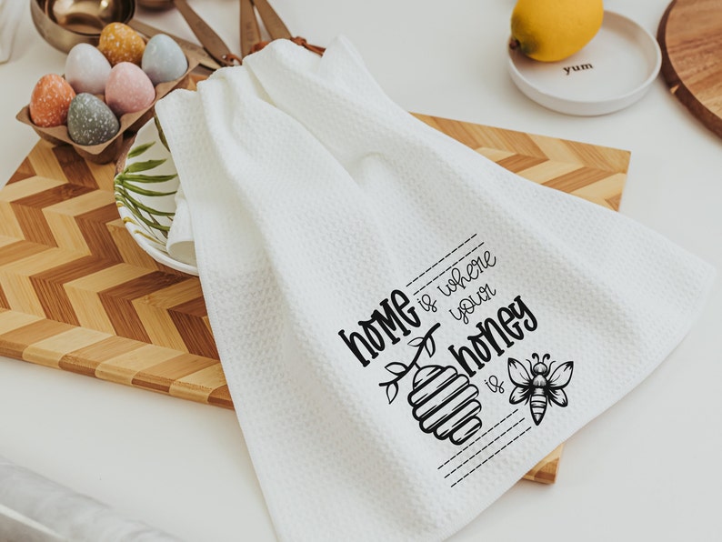 May include: White kitchen towel with black text that reads "Home is where your honey is" with a bee and honeycomb illustration.