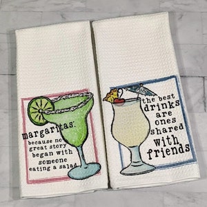 May include: Two white kitchen towels with colorful illustrations of cocktails and text. The first towel features a margarita with a lime wedge and the text "margaritas: because no great story began with someone eating a salad". The second towel features a tropical drink with a cherry and umbrella and the text "the best drinks are ones shared with friends".