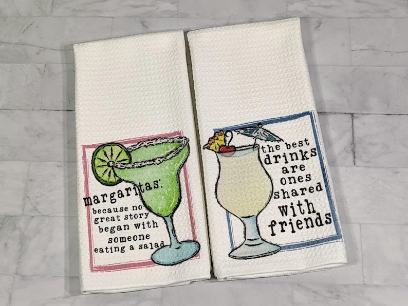 May include: Two white kitchen towels with colorful illustrations of cocktails and text. The first towel features a margarita with a lime wedge and the text "margaritas: because no great story began with someone eating a salad". The second towel features a tropical drink with a cherry and umbrella and the text "the best drinks are ones shared with friends".