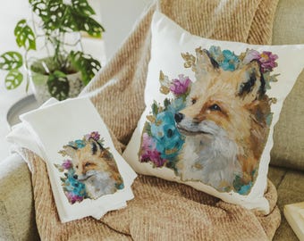 Op de afbeelding: Een wit kussen en een stapel witte handdoeken tonen een aquarel schilderij van een vos met blauwe, paarse en gouden bloemaccenten. De vos heeft bruine en bruine vacht. De items liggen op een beige deken.