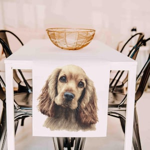 May include: A white table runner featuring a watercolor-style portrait of a Cocker Spaniel dog. The dog has long, wavy, light brown fur and dark eyes. The runner is on a white table with a gold-colored wire bowl.