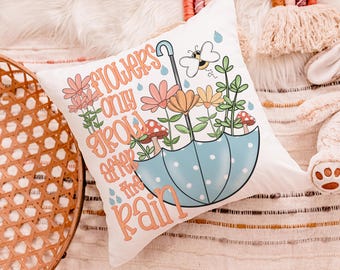 Op de afbeelding: Wit vierkant kussen met een grillig ontwerp met een paraplu met bloemen, paddenstoelen en een bij. De tekst luidt "Flowers only grow after the rain" in oranje letters. De paraplu is lichtblauw met witte stippen.