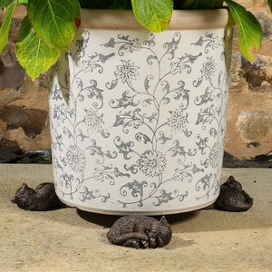 May include: Three small, sleeping cat figurines made of dark brown metal, placed around the base of a white ceramic planter with a grey floral pattern.