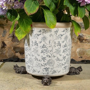 May include: A white ceramic planter with a blue floral pattern is surrounded by four small, gray metal cat figurines. The planter is filled with a green plant with purple flowers.