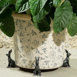 May include: A white ceramic planter with a floral pattern and three small silver rabbit figurines placed around the base.