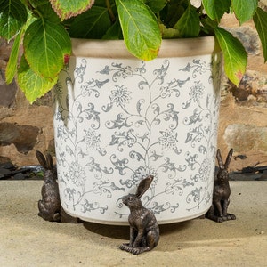 Op de afbeelding: Vier bronzenen konijnenbeeldjes zijn rond de basis van een witte keramische plantenpot met een bloemenpatroon geplaatst. De pot is gevuld met groen gebladerte.