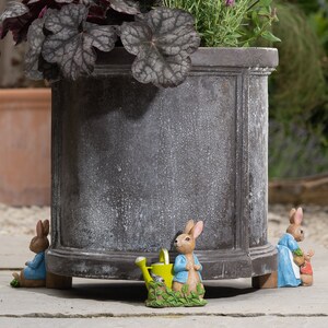 Op de afbeelding: Vier kleine keramische beeldjes van Peter Rabbit-karakters, waaronder Peter Rabbit met een gieter, zijn geplaatst rond de basis van een grote grijze plantenbak. De plantenbak is gevuld met groene planten.