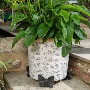 May include: A potted plant with pink gerbera daisies in a white ceramic planter with a blue floral pattern. The planter sits on a stone patio with decorative butterfly-shaped plant supports.