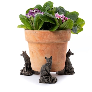 Könnte beinhalten: Ein Terrakotta-Blumentopf mit einer pink- und lilafarbenen Blühpflanze steht auf einem dekorativen Ständer mit vier Fuchsfiguren. Der Ständer ist aus dunkelbraunem Metall.