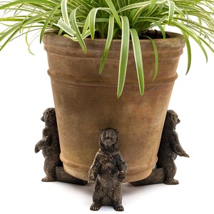 Op de afbeelding: Een terracotta plantenpot met een groene plant staat op een decoratieve standaard met drie bronzen berenfiguren. De beren zijn rond de basis van de pot geplaatst, wat een grillige en rustieke uitstraling creëert.