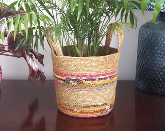 Puede incluir: Una planta de interior verde en una cesta tejida con rayas de colores. La cesta tiene dos asas.