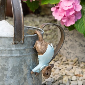 May include: A small brown and blue figurine of a mouse hanging upside down from the edge of a metal watering can. The mouse is wearing a blue shirt and has a brown tail.