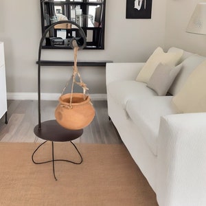 May include: A black metal plant stand with a curved arm and a hanging terracotta pot. The stand is on a brown rug in a living room with a white sofa.