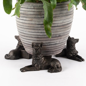 May include: A grey planter with a striped design is raised on three bronze dog figurines. The dogs are lying down with their heads facing forward.