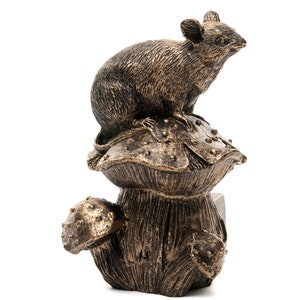 Op de afbeelding: Een bronzen beeldje van een muis die op een paddenstoel zit. De paddenstoel heeft een grote hoed en een kleinere paddenstoel die aan de zijkant groeit.