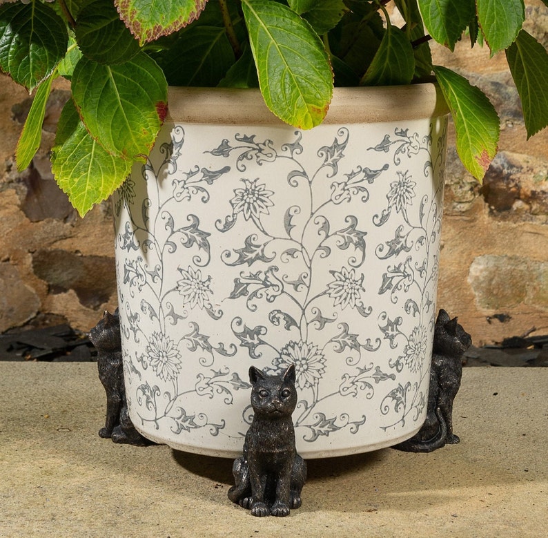 May include: A white ceramic planter with a black and white floral design. Four black cat figurines are used as plant pot feet.