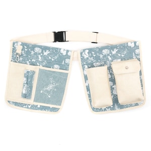 Può includere: Una cintura da giardinaggio in tela blu chiaro con più tasche e una stampa floreale bianca. La cintura ha una fibbia nera e un cinturino bianco.