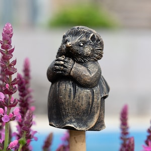 May include: A bronze-colored garden statue of a hedgehog wearing a dress. The hedgehog is standing on a wooden stake and has its paws clasped in front of it.