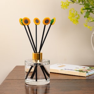 Pode incluir: Uma garrafa de difusor de vidro com tampa dourada e varetas difusoras pretas. As varetas são coroadas com pequenas decorações de girassol amarelas e marrons.