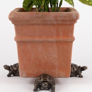 May include: A terracotta planter with a green plant is elevated on four bronze turtle figurines. The turtles are facing different directions and have detailed features.