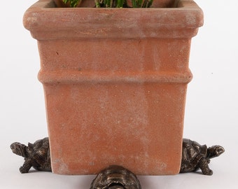 May include: A terracotta planter with a green plant is elevated on four bronze turtle figurines. The turtles are facing different directions and have detailed features.